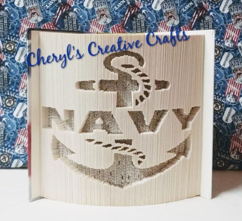 Puede incluir: Escultura de libro con la palabra "NAVY" y un ancla recortada en las p&aacute;ginas. El texto "Cheryl's Creative Crafts" est&aacute; escrito en azul en cursiva. El fondo tiene un patr&oacute;n patri&oacute;tico.