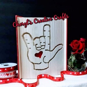 May include: A red book with the text "Cheryl's Creative Crafts" on the cover. The book is folded into a three-dimensional design of a hand making the sign for "I love you" in American Sign Language. A red heart is cut out of the center of the hand.