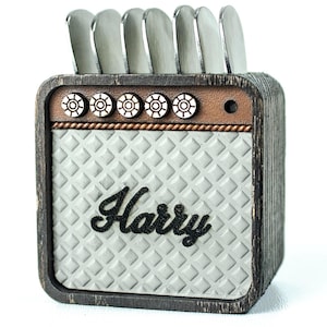 May include: A small, grey, wooden amplifier with a black speaker grille and the name "Harry" printed on the front. The amplifier has six knobs on the top and a brown wooden base.