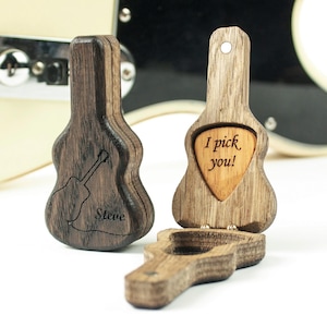 Könnte beinhalten: Holzgitarren-Plektrumhalter mit einer Gitarre im Hintergrund. Ein Halter hat ein Plektrum mit der Aufschrift "I pick you!" und ein anderer den Namen "Steve" eingraviert. Die Halter sind aus Holz.