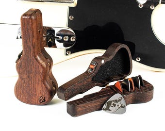 May include: A white electric guitar with a black pickguard positioned behind three small, dark brown wooden guitar-shaped cases. One case is closed, while two are open, revealing several guitar picks inside, including one with a cat illustration.