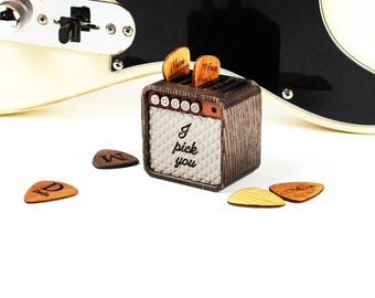 May include: A small wooden guitar pick holder shaped like a vintage amplifier with the text I pick you on the front. The holder contains two wooden guitar picks, with three additional wooden guitar picks scattered on the surface in front of it.