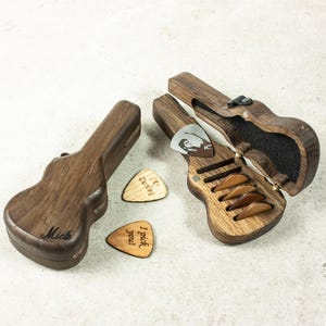 以下が含まれることがあります： ギターピックを収納するための木製のギター型の箱が2つ。1つの箱は閉まっており、「Mick」という名前が刻まれています。もう1つの箱は開いており、ピックを収納するためのスロットが6つあります。テーブルの上にはギターピックが3つあり、1つは「My Guitar」という文字が書かれており、1つは「I pick you!」という文字が書かれており、1つは白黒の人物の画像が描かれています。