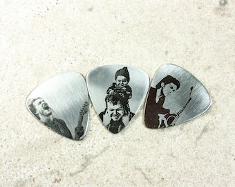 May include: Three silver-toned metal guitar picks featuring laser-engraved black and white photographic images. The left pick shows a musician playing guitar, the center pick depicts a child on shoulders, and the right pick shows a musician.