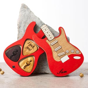 Könnte beinhalten: Roter Holz-Plektrumhalter mit drei Plektren. Der Halter ist mit den Worten "I pick you!" graviert. Der Plektrenhalter befindet sich neben einer roten Holzgitarre mit dem Namen "Sam" darauf.