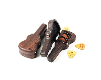 May include: Two dark brown wooden guitar-shaped cases, one closed and one open, revealing a black interior lining. The open case holds several guitar picks. Two additional light-colored wooden guitar picks with engraved text lie nearby.
