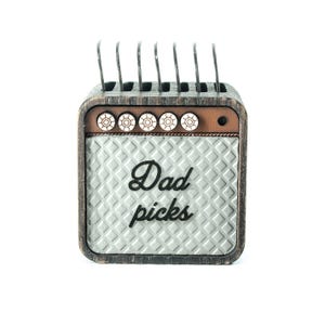 Könnte beinhalten: Ein Gitarren-Plektrumhalter aus Holz in Form eines Vintage-Verstärkers mit dem Text "Dad picks" auf der Vorderseite. Der Halter hat eine braune Holzoptik und ein silbernes Metallgitter. Auf der Oberseite des Halters befinden sich sechs Knöpfe.