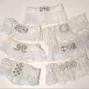 Op de afbeelding: Collectie witte en lichtblauwe bruidsgarterriemen met diverse strass versieringen. De jarretels hebben kant- en tule details, met ontwerpen zoals strikken, bloempatronen en sprankelende kristallen accenten. Deze accessoires zijn ontworpen voor bruiloften en speciale gelegenheden.
