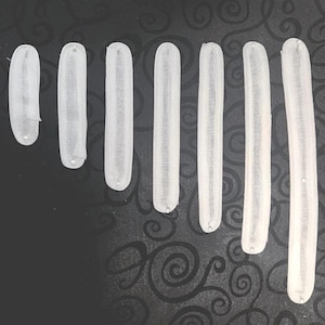 May include: Seven white, elongated, semi-sheer fabric strips of varying lengths arranged vertically. A metal ruler is positioned on the left, showing the shortest strip is approximately 2.5 inches and the longest is approximately 7.5 inches long.