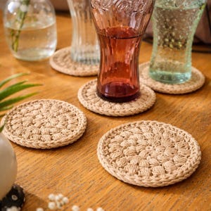 May include: A collection of beige crochet coasters, each with a circular design, are arranged on a wooden table. Several Coca-Cola glasses, some filled with liquid, rest on the coasters. A small vase with white flowers is in the background.