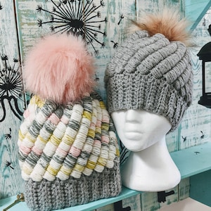 Puede incluir: Dos gorros de punto de ganchillo grises con diferentes patrones y pompones. Un gorro tiene un pompón rosa y un patrón a rayas blanco, gris y amarillo. El otro gorro tiene un pompón marrón y un patrón gris texturizado.