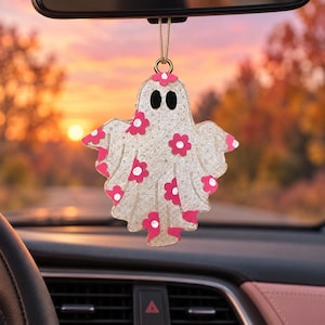 May include: A ghost-shaped car air freshener or hanging ornament featuring a translucent, glittery white finish decorated with bright pink flower patterns. The ghost has two black oval eyes and is suspended by a thin gold-colored string.