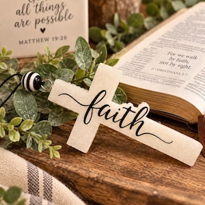 May include: A white, textured cross ornament featuring the word faith in black cursive script. The cross is attached to a black cord with a black and white striped bead. The design is simple, clean, and focuses on the central religious symbol.