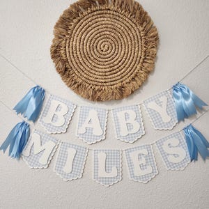 May include: A baby shower banner with the name "Miles" in white letters on blue gingham flags, accented with blue ribbons. A round woven wall hanging with a fringed edge is above the banner.