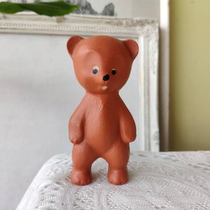 May include: A brown ceramic teddy bear figurine with black painted eyes and a small black nose. The bear is standing upright with its arms at its sides.