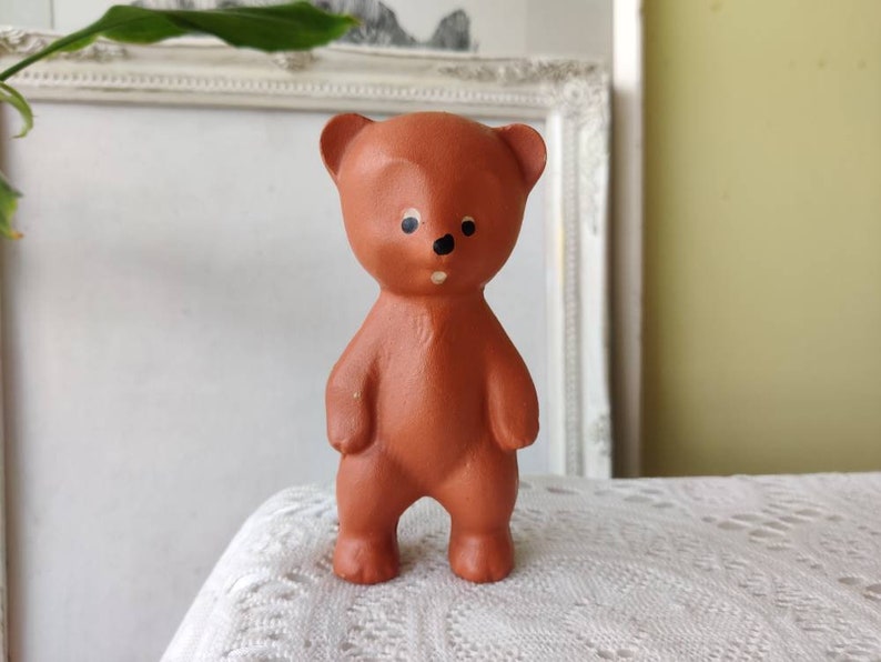 May include: A brown ceramic teddy bear figurine with black painted eyes and a small black nose. The bear is standing upright with its arms at its sides.
