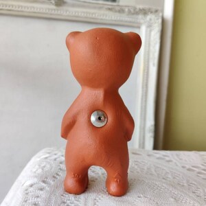 May include: A terracotta bear figurine with a silver button on its back. The bear is standing on a white surface.