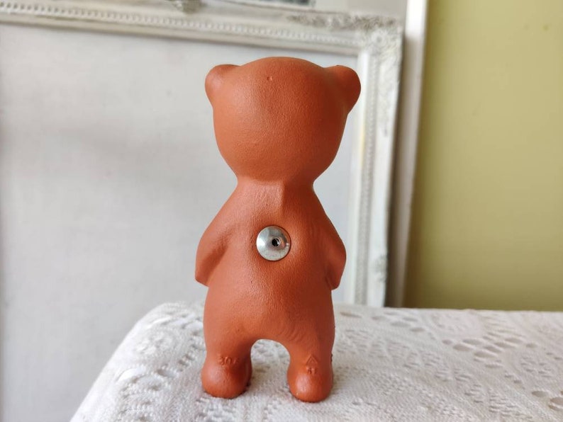 May include: A terracotta bear figurine with a silver button on its back. The bear is standing on a white surface.