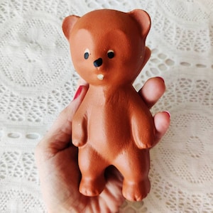 May include: A brown rubber toy bear with black eyes and a black nose. The bear is standing upright with its arms at its sides.