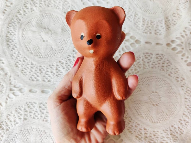 May include: A brown rubber toy bear with black eyes and a black nose. The bear is standing upright with its arms at its sides.