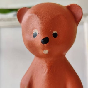 May include: A terracotta-coloured ceramic bear figurine with black painted eyes and a black nose. The bear has a white painted mouth and a slightly surprised expression.