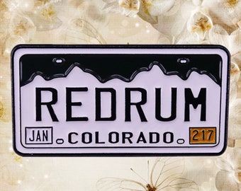 Puede incluir: Un pin de esmalte negro y blanco con forma de matrícula con la palabra "REDRUM" en letras negras sobre fondo blanco. La matrícula también tiene las palabras "JAN" y "COLORADO" en letras negras sobre fondo blanco, y el número "217" en letras naranjas sobre fondo blanco.