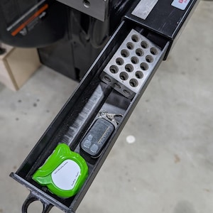 May include: A black plastic storage tray attached to a table saw fence, containing a silver metal 1-2-3 block with circular holes, a black Laguna remote control, and a bright green FastCap measuring tape with the text Lefty / Righty.