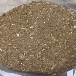 May include: A close-up view of a bag filled with soil. The soil is a mix of brown and gray tones, with small white and tan particles scattered throughout. The texture appears granular and slightly coarse, suggesting a potting mix.