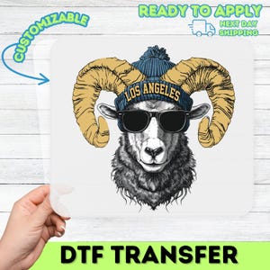 Peut inclure: Transfert DTF représentant un bélier portant des lunettes de soleil et un bonnet bleu avec le texte "LOS ANGELES". Le bélier a de grandes cornes beiges et un visage noir et blanc. Le transfert est sur fond blanc avec texte "PERSONNALISABLE" et "PRÊT À APPLIQUER".