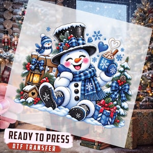 May include: A DTF transfer sheet featuring a cheerful snowman wearing a black top hat and blue plaid scarf, holding a mug of hot cocoa. The design includes a blue bird, a lantern, a gift box, and pine branches with red berries and snowflakes.