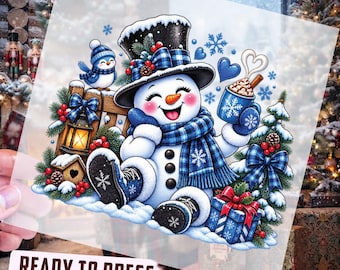 Könnte beinhalten: Ein DTF-Transferbogen mit einem fröhlichen Schneemann, der einen schwarzen Zylinder und einen blau karierten Schal trägt und eine Tasse heiße Schokolade hält. Das Design umfasst einen blauen Vogel, eine Laterne, ein Geschenk und Tannenzweige mit Beeren.