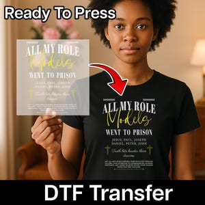 May include: A black t-shirt featuring white and yellow text that reads: ALL MY ROLE Models WENT TO PRISON, JESUS, PAUL, JOSEPH, DANIEL, PETER, JOHN, Truth hits harder than dreams, NOT ALL INFLUENCE INSPIRES. SOME EDUCATES THROUGH FAILURE.