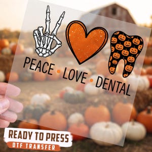 May include: A clear DTF transfer sheet featuring a graphic with a skeleton hand making a peace sign, an orange heart, and a tooth filled with a jack-o'-lantern pattern. Below the icons, the text reads PEACE LOVE DENTAL in black capital letters.
