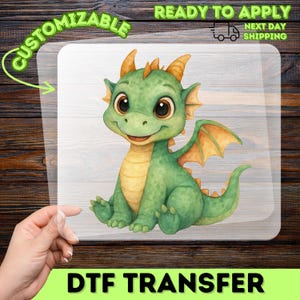 May include: A DTF transfer featuring a cartoon dragon. The dragon is green with yellow accents, large brown eyes, and orange wings. The words "CUSTOMIZABLE" and "READY TO APPLY" are printed on the transfer. The words "DTF TRANSFER" are printed at the bottom.