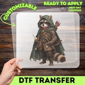 May include: A clear transfer sheet featuring a graphic illustration of a raccoon dressed as a fantasy archer. The raccoon wears a dark green hooded cloak, brown leather armor, and carries a bow, arrows, and a small dagger at its side.