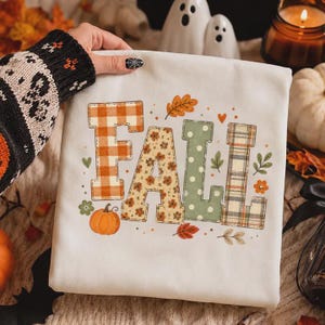 Peut inclure: Un sweat-shirt blanc avec le mot FALL écrit en grandes lettres majuscules. Chaque lettre présente un motif distinct : vichy orange, fleurs, pois vert sauge et tartan. Le design inclut des petites citrouilles orange et des feuilles.