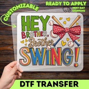 May include: A clear DTF transfer sheet featuring a baseball-themed graphic with the text HEY BROTHER, Brother SWING! The design includes two crossed baseball bats, a large red polka-dot bow, several baseballs, and small yellow hearts.