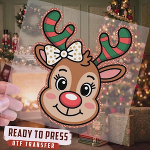 May include: A clear DTF transfer sheet featuring a cartoon reindeer head with a red nose, pink cheeks, and a white bow with gold polka dots. The reindeer has green and red striped antlers outlined in a copper glitter effect. Text reads READY TO PRESS.