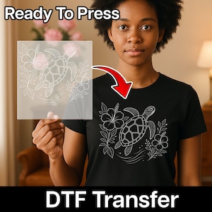 May include: A black t-shirt featuring a white line art graphic of a sea turtle surrounded by hibiscus flowers and leaves. A transparent sheet displays the same white sea turtle and floral design, indicating a ready to press DTF transfer.