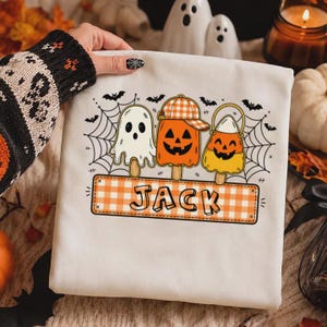 May include: A white sweatshirt featuring a graphic of three Halloween-themed popsicles: a white ghost, an orange jack-o'-lantern wearing a plaid cap, and a candy corn character. Below them is an orange plaid rectangular patch with the name JACK.