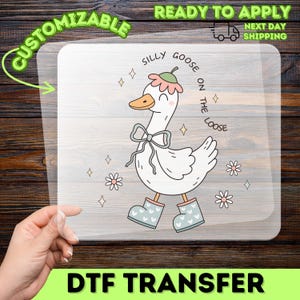 May include: A DTF transfer featuring a cartoon goose with the text "Silly Goose on the Loose." The goose is white with a pink and green flower hat, blue boots, and a blue bow. The transfer is customizable and ready to apply.