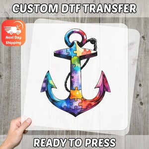 Puede incluir: Transferencia de diseño de ancla colorida con un patrón de piezas de rompecabezas. El ancla es en tonos de azul, morado, amarillo y rojo. Se muestra el texto "CUSTOM DTF TRANSFER" y "READY TO PRESS". La imagen también incluye un icono de "Next Day Shipping".