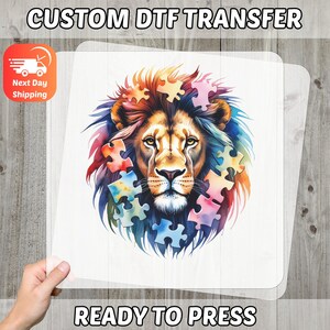 May include: A ready-to-press DTF transfer featuring a colorful lion's face with puzzle pieces. The lion has a brown mane and is surrounded by puzzle pieces in various colors. The text "CUSTOM DTF TRANSFER" and "READY TO PRESS" are displayed.