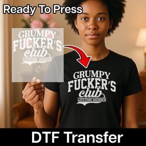 May include: A black t-shirt featuring the white text GRUMPY FUCKER'S club LIFETIME MEMBER. A transparent sheet displays the same white graphic design, indicating a DTF transfer. A red arrow points from the transfer sheet to the t-shirt design.