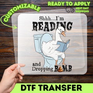 May include: A clear DTF transfer sheet featuring a cartoon white goose wearing glasses, sitting on a light blue toilet while reading a blue book. The text reads Shhh...I'm READING and Dropping BOMB with a brown poop emoji replacing the letter O.