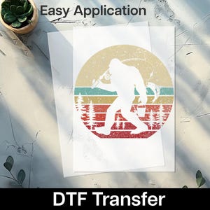 May include: White DTF transfer featuring a Bigfoot silhouette fishing, set against a retro sunset design with red, yellow, and teal stripes. The words "Easy Application" and "DTF Transfer" are printed on the image.