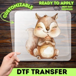 May include: A DTF transfer featuring a watercolor illustration of a smiling squirrel holding an acorn. The squirrel has brown fur, a fluffy tail, and rosy cheeks. The words "CUSTOMIZABLE" and "READY TO APPLY" are printed on the transfer.
