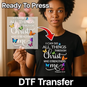 Op de afbeelding: Zwart T-shirt met de tekst "Ik kan alles doen door Christus die mij kracht geeft" en vlindergraphics. Een heldere DTF-transfer met hetzelfde ontwerp wordt naast het shirt gehouden. De woorden "Ready To Press" en "DTF Transfer" zijn ook zichtbaar.