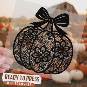 May include: A black DTF transfer featuring a pumpkin silhouette with a detailed lace and floral pattern, topped with a large bow. The design is printed on a clear film sheet with the text READY TO PRESS DTF TRANSFER printed at the bottom.
