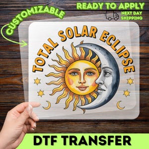 Può includere: Un trasferimento DTF con un disegno di sole e luna e la scritta "TOTAL SOLAR ECLIPSE". Il sole è giallo e arancione con un volto sorridente, e la luna è argentata. Il trasferimento è su un supporto trasparente.
